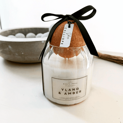 A candle with a label reading 'Ylang & Amber' placed on a table with a black bow on top, indicating it is a hand-poured soy candle.