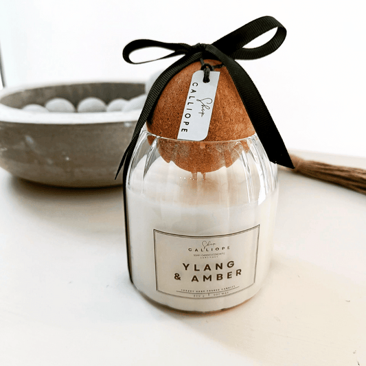 A candle with a label reading 'Ylang & Amber' placed on a table with a black bow on top, indicating it is a hand-poured soy candle.