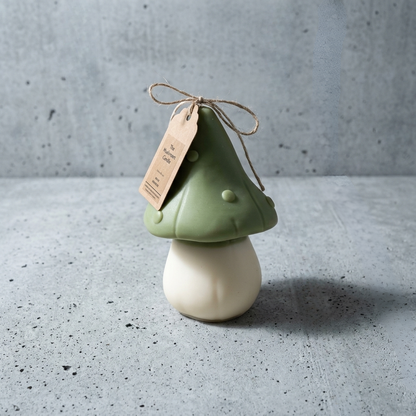 A small mushroom-shaped decorative object with a sage green cap and an off-white rounded base, tied with twine and a kraft paper tag, set on a gray speckled surface against a gray wall.