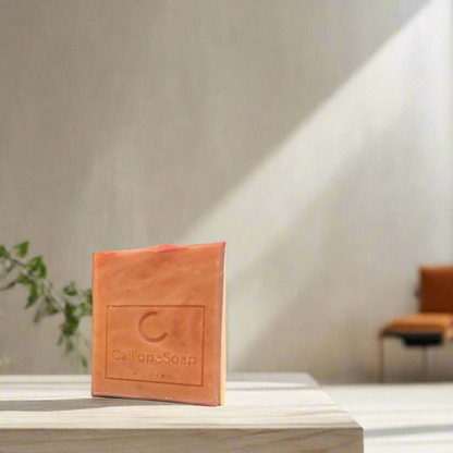 A bar of handmade soap with red clay, branded with 'calliopeshop.com', placed on a wooden surface with a plant in the background.