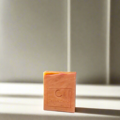 A bar of handmade soap with red clay, branded with 'calliopeshop.com', placed on a wooden surface with a plant in the background.