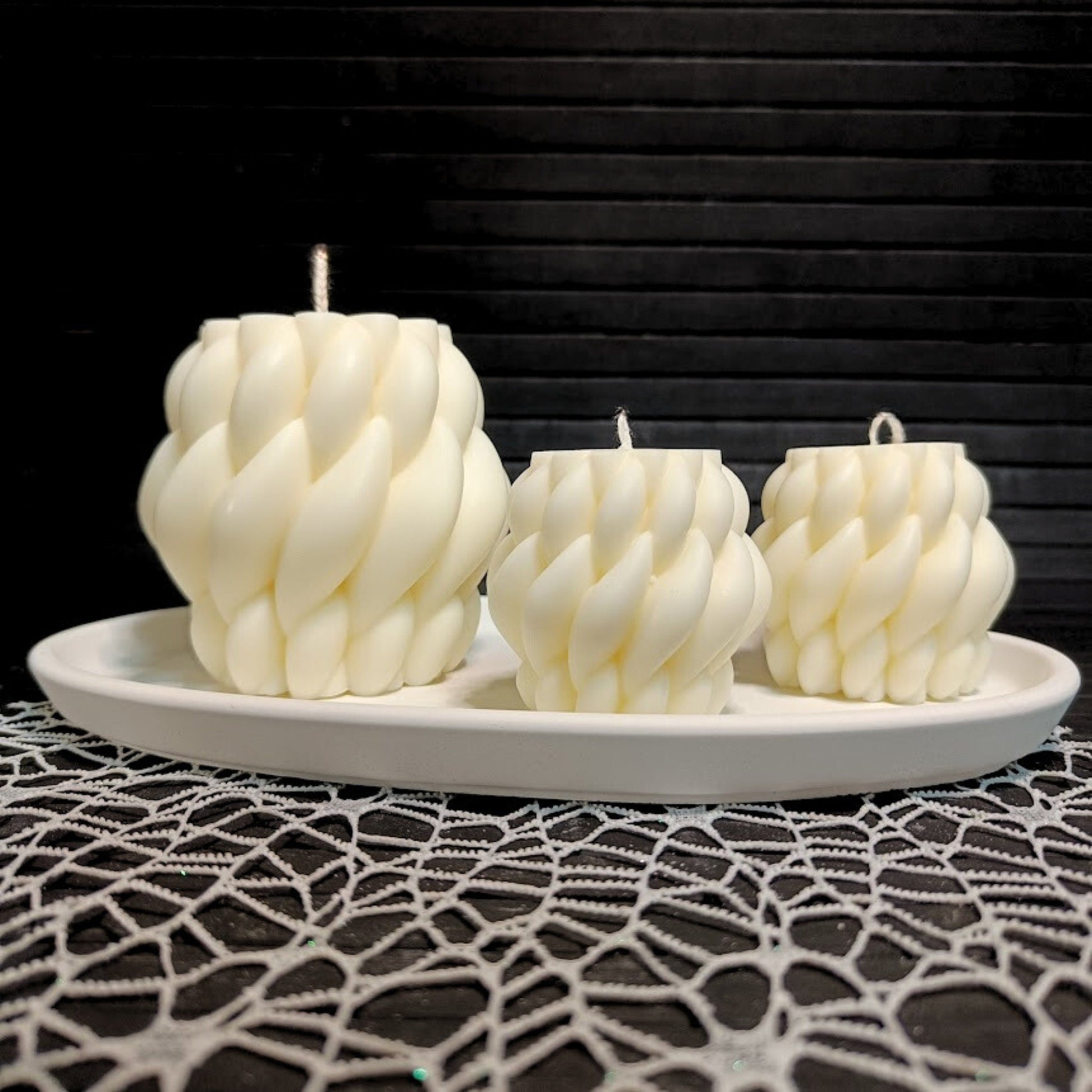 Three off-white soy wax pillar candles with a twist design, displayed on a round white plate, with a dark textured background.