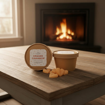 Two containers of 'Orange Cinnamon' candle on a wooden surface with a fireplace in the background.