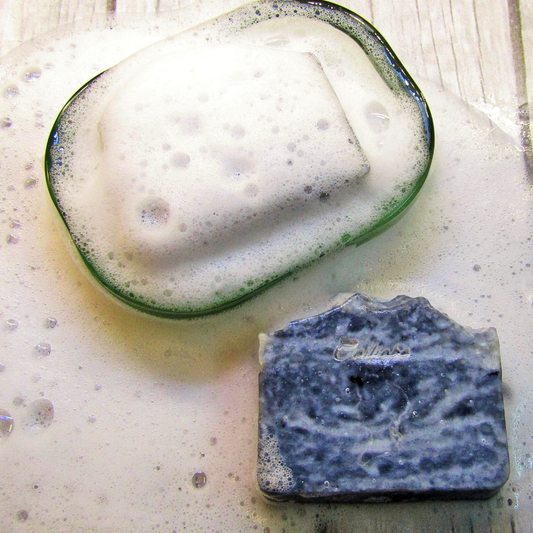 Two soap bars, one with a green rim and white foam, the other blue and textured, on a light wooden surface.