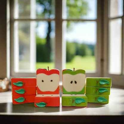Handmade apple-shaped soap bars with green and red leaves on a window sill, with a blurred background.