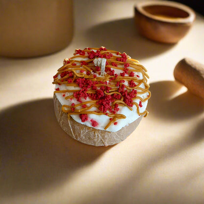 A decorative candle designed to resemble a red velvet cake with white icing and red sprinkles, set on a wooden surface.