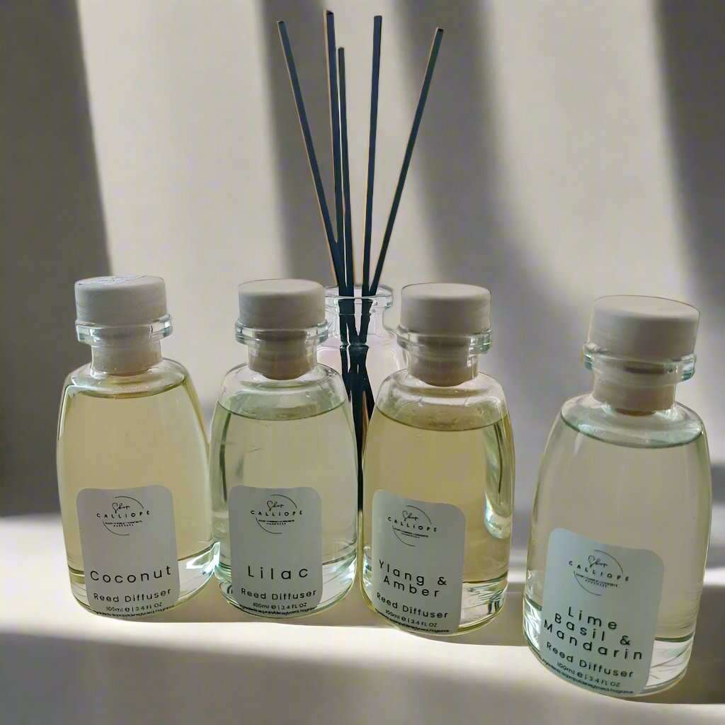 A collection of four reed diffuser bottles with clear labels and white reeds. The bottles are labeled with different scents: coconut, lilac, amber, and rose.
