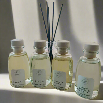 A collection of four reed diffuser bottles with clear labels and white reeds. The bottles are labeled with different scents: coconut, lilac, amber, and rose.