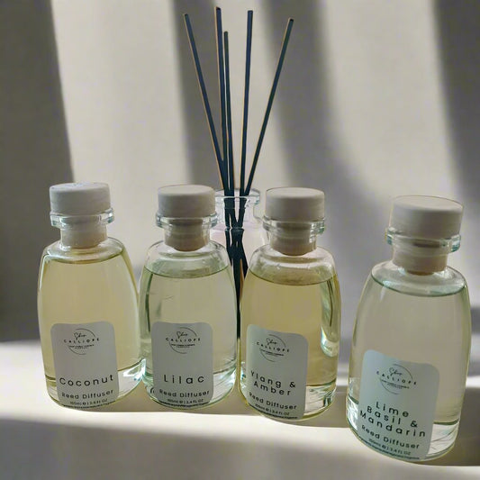 A collection of four reed diffuser bottles with clear labels and white reeds. The bottles are labeled with different scents: coconut, lilac, amber, and rose.