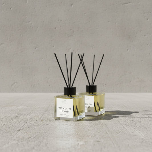 A set of clear glass reed diffuser bottles with black caps and white labels, arranged on a light wood shelf. The front row includes 'Welcome Home', 'Vanilla Fresh', and 'Clean & Fresh' scents, with more bottles stacked behind them.