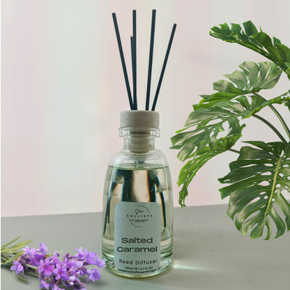 Glass diffuser bottle labeled 'Salted Caramel' with black sticks on a light surface with green leaves and purple flowers in the background.