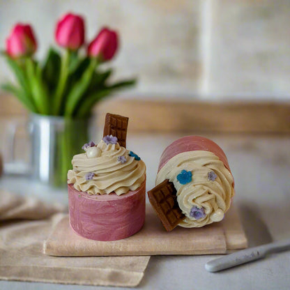 Handmade soap shaped and decorated to resemble a cupcake with pink layers, white frosting, and chocolate pieces, set against a neutral background.