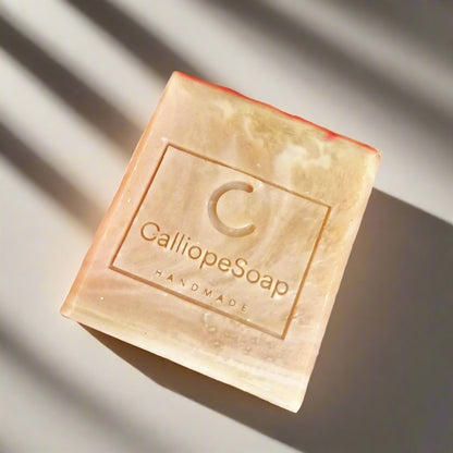 A bar of handmade soap with red clay, branded with 'calliopeshop.com', placed on a wooden surface with a plant in the background.