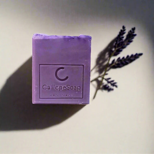 A bar of lavender-scented soap with 'CALLIOPE SOAP' branding, placed on a surface with some lavender flowers in the background.