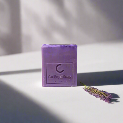 A bar of lavender-scented soap with 'CALLIOPE SOAP' branding, placed on a surface with some lavender flowers in the background.