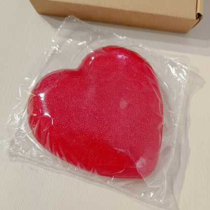 Red heart-shaped object in clear plastic packaging on a beige surface with a cardboard box in the background.