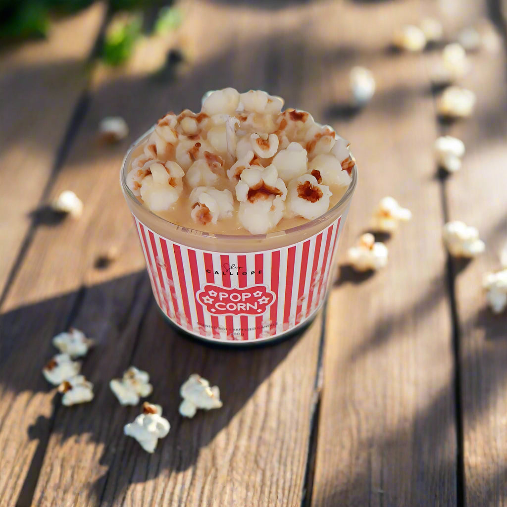 A handmade soy wax candle in a glass container designed to resemble a bucket of popcorn, with scattered popcorn pieces around it.
