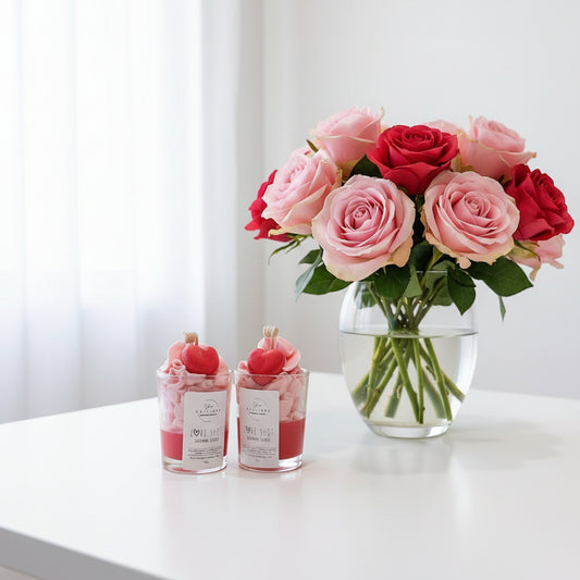 Bouquet of pink and red roses in a clear vase on a white surface with two love shot candles.