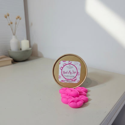 Pink lip-shaped wax melts in front of a round container with a label on a light surface.