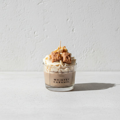 Ένα κερί σε γυάλινο ποτήρι με την ετικέτα 'WHISKEY CARAMEL' από CalliopeShop, γεμισμένο με κρεμώδες μπεζ κερί και διακοσμημένο με  κερί σε σχήμα και χρώμα καραμέλας, τοποθετημένο σε μια ανοιχτόχρωμη επιφάνεια με σκούρο φόντο.