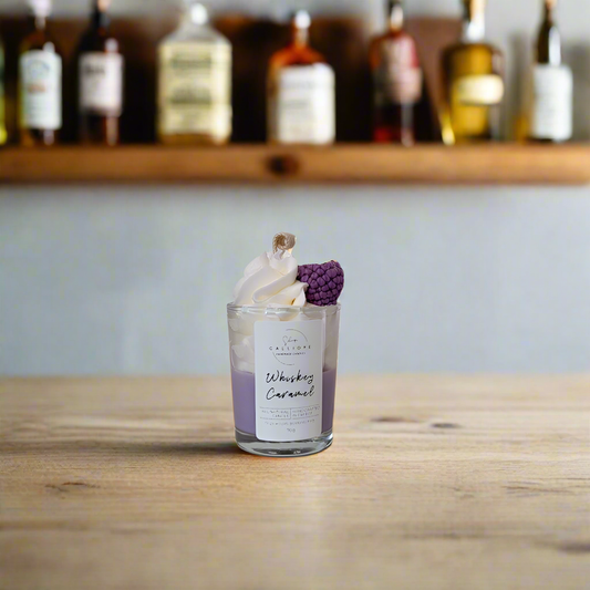 Candle labeled 'Whiskey Caramel' with decorative elements on a wooden surface, with bottles in the background.
