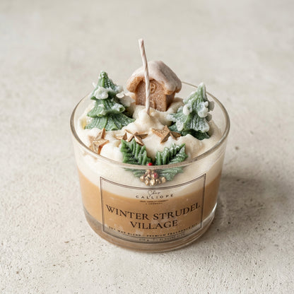 A glass jar candle labeled 'WINTER STRUDELL VILLAGE' by Calliope, filled with layered beige and white wax and topped with miniature green Christmas trees, a gingerbread house, and a white wick, set on a light surface with a blurred indoor background.